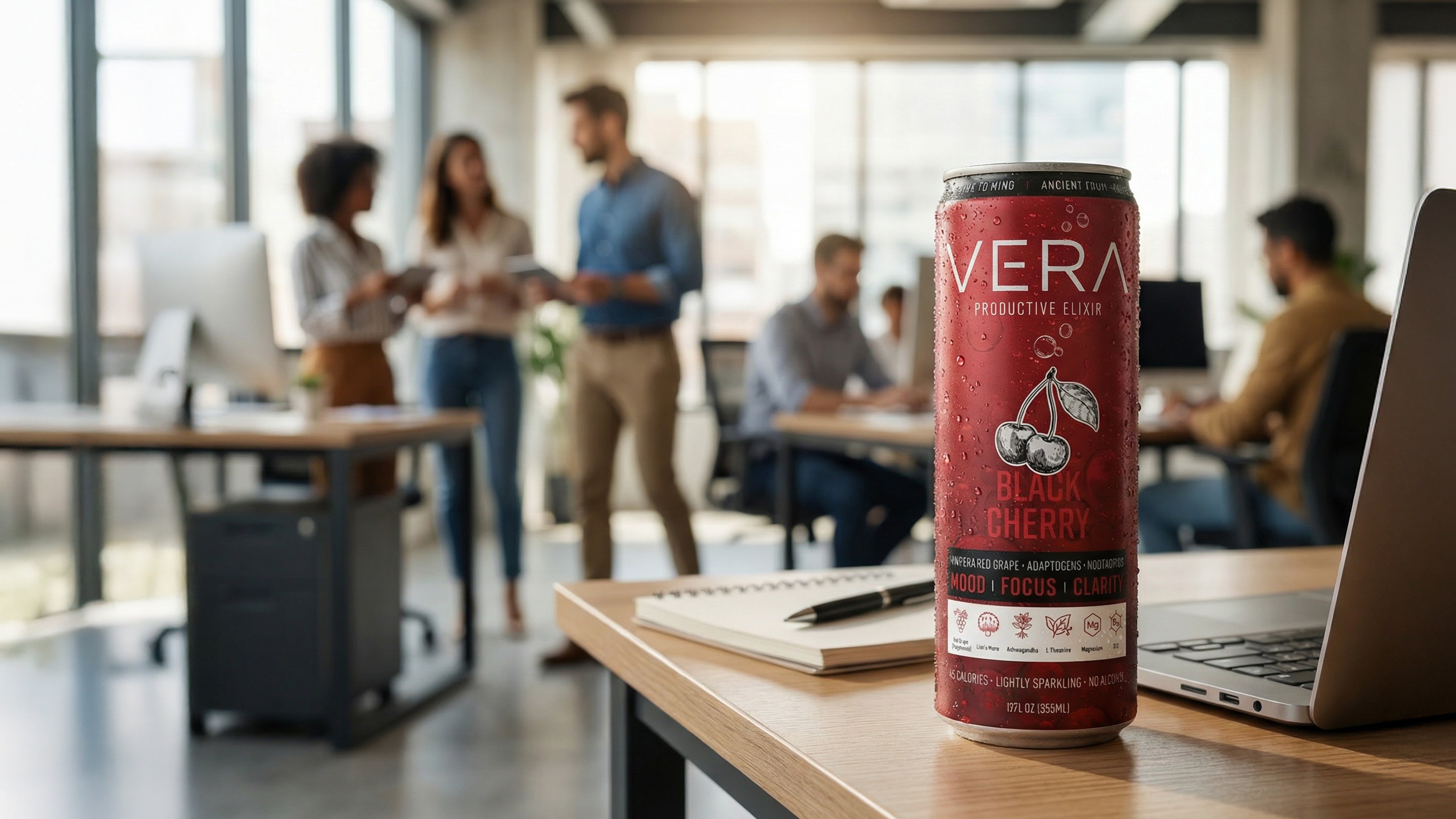 A can of Vera Black Cherry Productive Elixir stands on a wooden desk in a modern, sunlit office with coworkers collaborating in the blurred background.